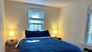 A bedroom with a large bed, two bedside tables with lamps, a window with blinds, and a radiator.