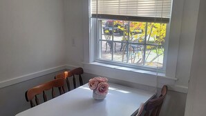Un comedor con una mesa blanca y sillas de madera, un jarrón con flores y una ventana con persianas.