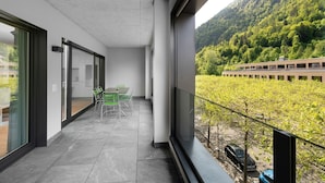 A balcony with a view of a forested hillside, a dining area with chairs, and a car parked outside.