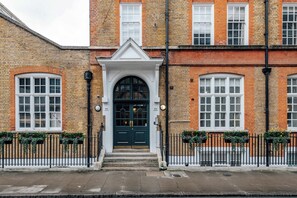 A traditional brick building with a central entrance, white-framed windows, and a black metal fence with flower boxes.