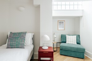 A bedroom with a bed, a red nightstand, a white lamp, a green sofa, and a window with blinds. - The Regency by nestor | 2BD LG Apartment (London)
