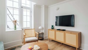 Een moderne woonkamer met een groot raam, een aan de muur bevestigde televisie, een houten kast en een ronde salontafel met lekkernijen.