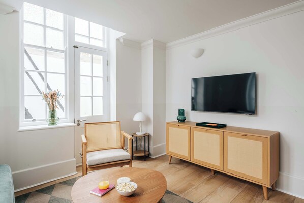 A modern living room with a large window, a television mounted on the wall, a wooden cabinet, and a round coffee table with snacks.