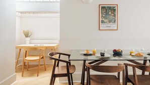 Een eetgedeelte met een glazen tafel, houten stoelen en een witte muur met een ingelijst beeld.
