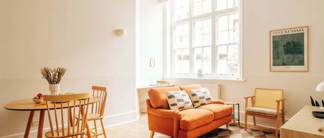 Ein helles Zimmer mit großem Fenster, einem Holzespeisetisch mit Stühlen, einer Couch und einem kleinen Beistelltisch.