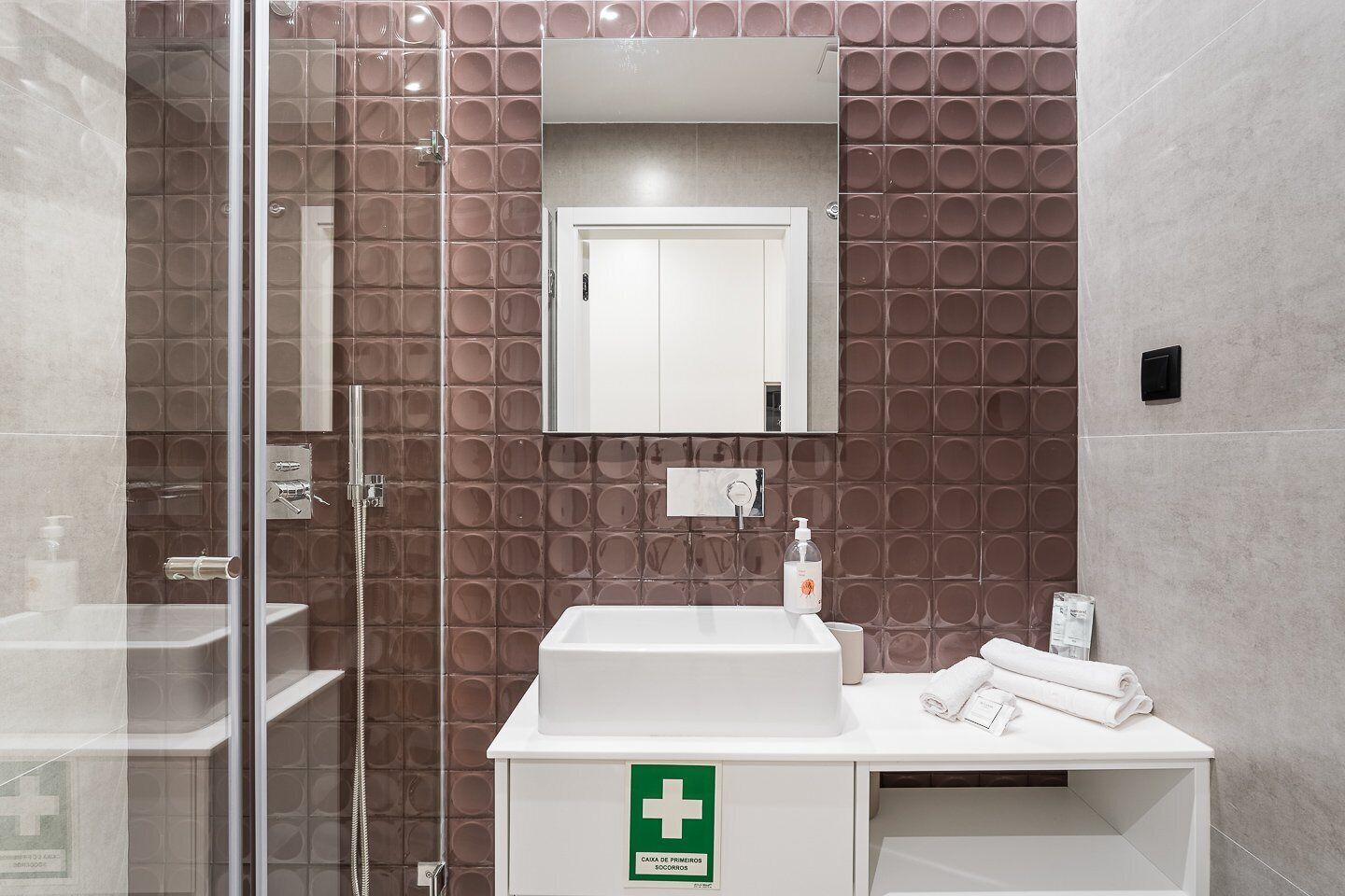 A modern bathroom with a glass shower enclosure, a white sink, and a mirror above it. There is a green emergency sign on the wall.