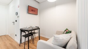 A compact room with a dining table set for two, a sofa with a decorative pillow, a mounted TV displaying "GUEST READY", and a ceiling light.