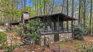 A rustic cabin with a stone chimney, a covered porch with chairs, and a stone staircase leading up to the entrance. - Highlands Close-to-Town Cabin, Pet-Friendly (Highlands)