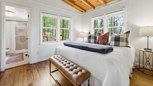 A bedroom with a wooden ceiling, large windows, a bed with multiple pillows, a bench, and a bedside table with a lamp. - Highlands Close-to-Town Cabin, Pet-Friendly (Highlands)