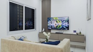 Ein modernes Wohnzimmer mit einem großen Flachbildfernseher an der Wand, einem beigefarbenen Sofa, einem Fenster mit Jalousien und einer dekorativen Vase mit Blumen auf einem Beistelltisch.