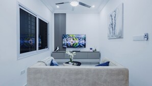 Ein modernes Wohnzimmer mit einem großen Flachbildfernseher, einem Deckenventilator und einem grauen Sofa mit blauen Kissen.