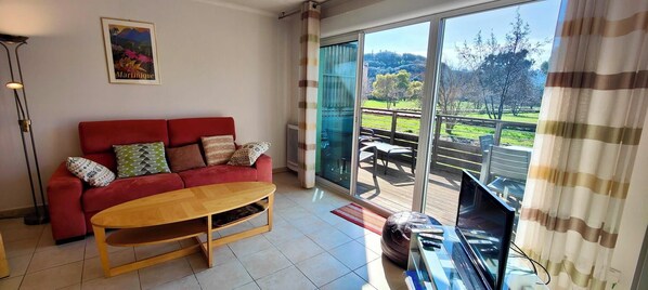 A living room with a red sofa, wooden coffee table, and a large sliding glass door leading to a balcony with a view of a green landscape. - Spacious apartment with pool (Saint-Raphaël)