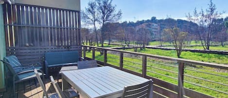 A balcony with a table and chairs, a bench, and a view of a green field and trees.