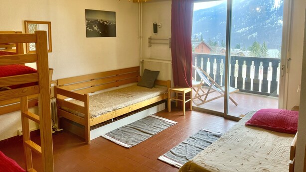 A dormitory room with bunk beds, a single bed, a small table, a chair, a radiator, a window with a view of a mountain, and a hanging light fixture. - Lovely T3 with loggias close to the slopes (Saint-Chaffrey)