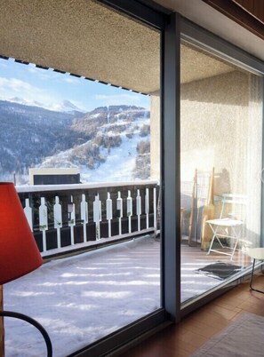 Un balcon offrant une vue sur des montagnes enneigées, une chaise rouge et une porte vitrée donnant sur une pièce où se trouvent une chaise blanche et une petite table.