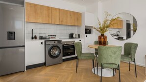A modern kitchen with wooden cabinets, a stainless steel refrigerator, a washing machine, and a dining area with green chairs and a round table. - Ratanga Park Escape | Century City (Cape Town)