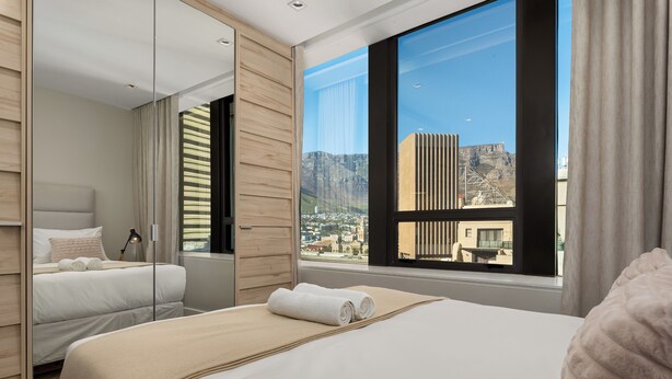 A hotel room with a large window, a bed with a beige blanket, a bedside table, and a mirror. - Modern Foreshore Apartment | Mountain Views (Cape Town)