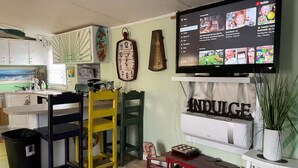 Un espacio habitable compacto con kitchenette, televisor montado en la pared y una pequeña zona de estar con un banco y sillas.