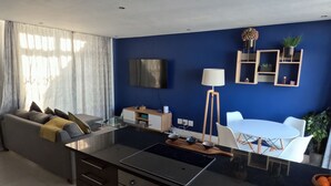 A modern kitchen with a central island, a dining area with white chairs, a sofa with cushions, and a flat-screen TV mounted on the wall. - Two Bedroom Beachfront Penthouse with Parking (Cape Town)