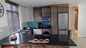 A modern kitchen with a central island, wooden stools, a coffee machine, and a refrigerator. - Two Bedroom Beachfront Penthouse with Parking (Cape Town)