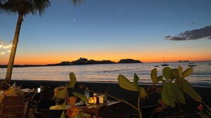 A beachside restaurant with tables and chairs, overlooking the sea and a sunset.