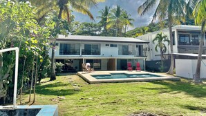 A modern two-story house with a swimming pool, surrounded by lush greenery and palm trees.