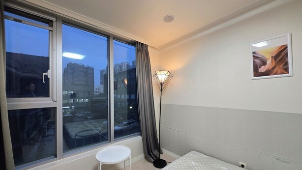 A bedroom with a large window, a standing lamp, a small table, and a bed.