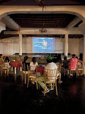 A group of people seated in a semi-circular arrangement facing a large screen in a room with a high ceiling and exposed beams.