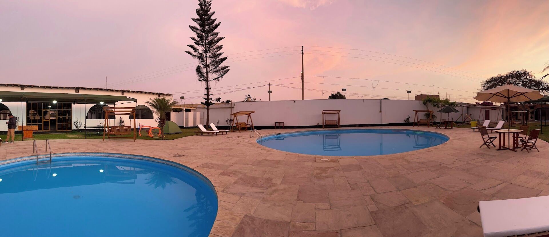 Un área de piscina con dos piscinas, sillones de descanso y un edificio con un porche cubierto.