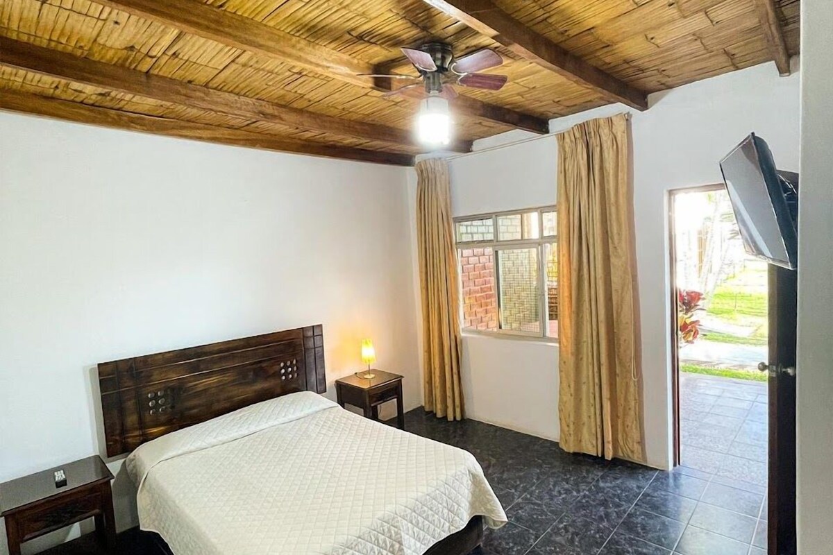 A room with a bed, a nightstand, a ceiling fan, a window with curtains, and a TV.