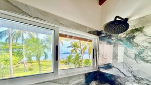 A bathroom with two windows offering views of a tropical landscape, a shower area, and a marble wall. - 37 steps to Playa Potrero from the back door. Pool, Sleeps 6! Sunsets galore! (Guanacaste Province)