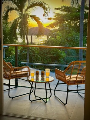 Un balcon avec des fauteuils en osier et une table, deux verres de bière et une vue sur un paysage tropical.