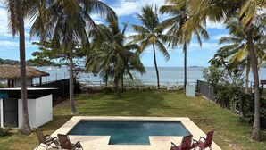 A rectangular swimming pool with two lounge chairs on each side, surrounded by palm trees and a view of the ocean. - 37 steps to Playa Potrero from the back door. Pool, Sleeps 6! Sunsets galore! (Guanacaste Province)