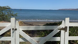 A beach view with a wooden gate, calm sea, and distant land. - 37 steps to Playa Potrero from the back door. Pool, Sleeps 6! Sunsets galore! (Guanacaste Province)