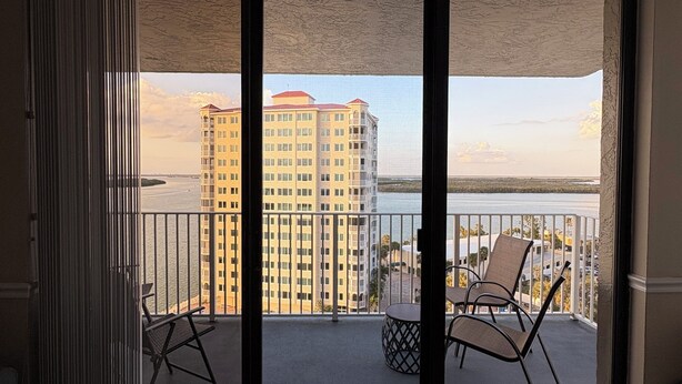 A balcony with two chairs and a small table, overlooking a body of water and high-rise buildings. - Lovers’ Key Beach Club by Check In Vacation Rental - 1 Bedroom Apartment #1005 (Bonita Springs)