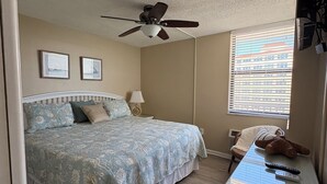 A bedroom with a bed, a ceiling fan, a window with blinds, a chair, and a desk with a teddy bear and remote controls. - Lovers’ Key Beach Club by Check In Vacation Rental - 1 Bedroom Apartment #1005 (Bonita Springs)