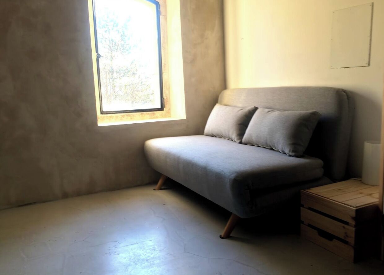 A small, dimly lit room with a single window, a grey sofa, and a wooden crate.