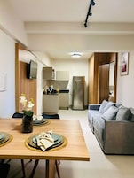 Cocina moderna con refrigerador, estufa y gabinetes. Hay un comedor con una mesa preparada para dos personas, un sofá y un televisor montado en la pared.