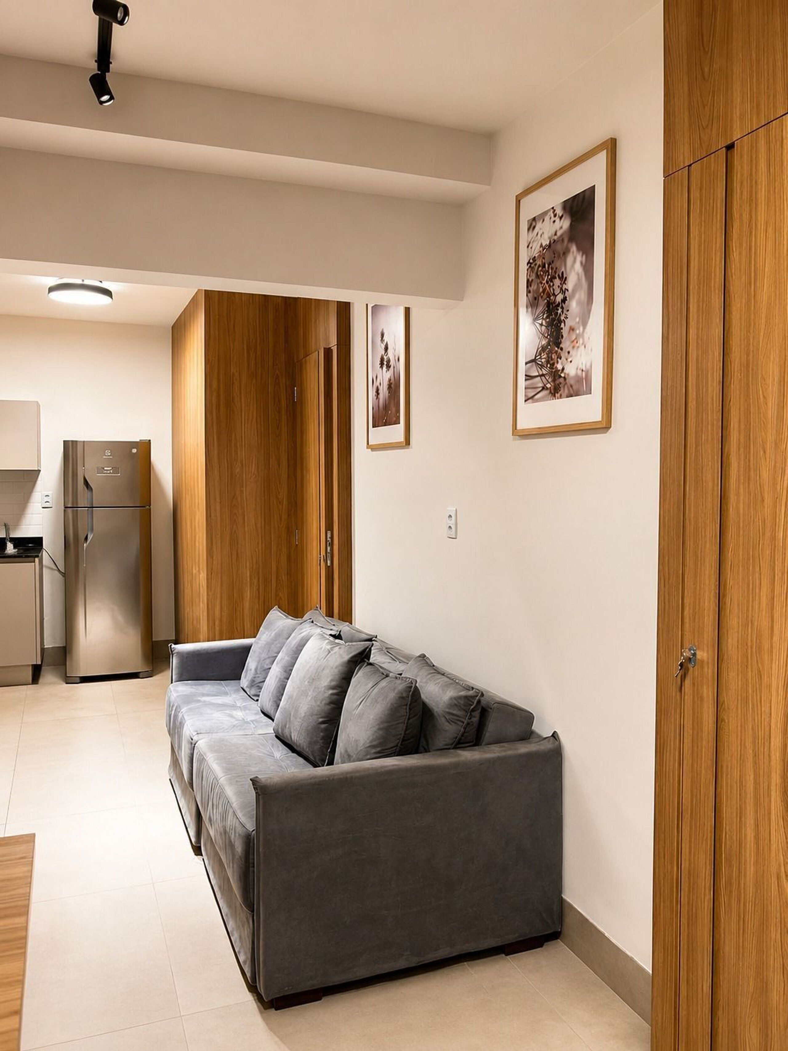 A modern living space with a grey sofa, wooden wardrobe, and a kitchen area with a refrigerator.
