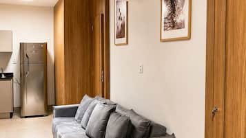 A modern living space with a grey sofa, wooden wardrobe, and a kitchen area with a refrigerator.