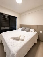 Un dormitorio con una cama grande, cabecera, ventana con persianas y una lámpara en el techo.