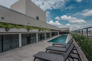 Un área de piscina en la azotea con vistas despejadas del horizonte urbano, arquitectura moderna y un espacio de descanso con sillones.