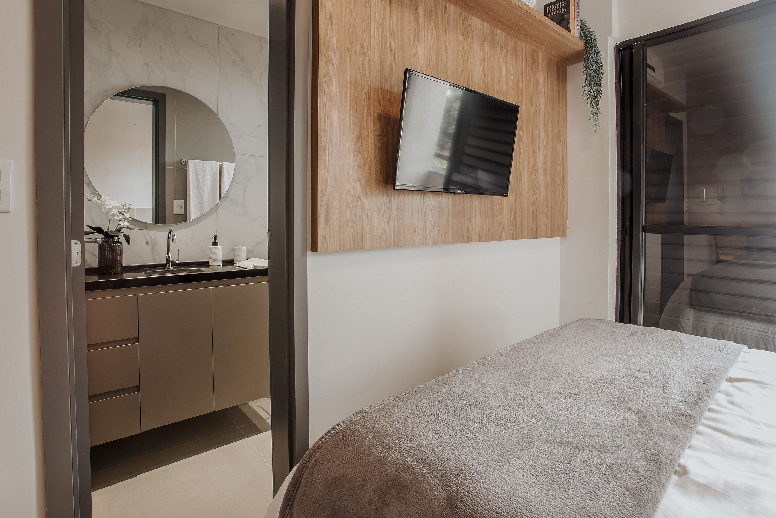 A modern bedroom with a bed, a TV mounted on a wooden panel, a bathroom with a marble wall, and a large window.