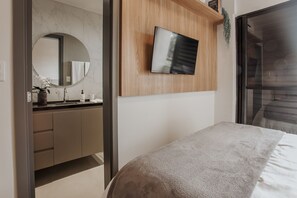 Un dormitorio moderno con cama, televisor montado en un panel de madera, baño con pared de mármol y un ventanal grande.