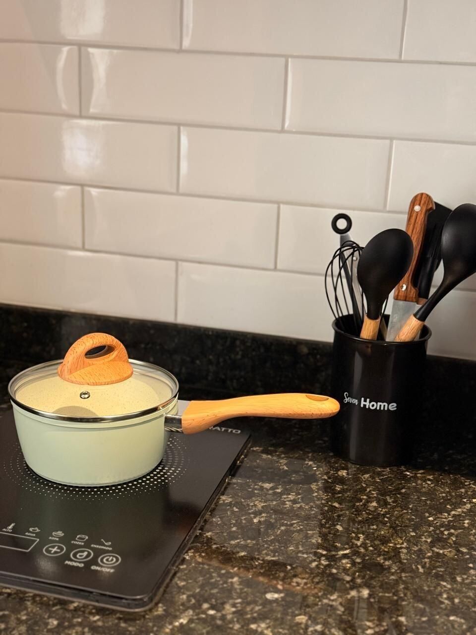 Una cocina con una olla de vidrio sobre una cocina de inducción, una cuchara de mango de madera y un portalapices con utensilios negros y de madera.