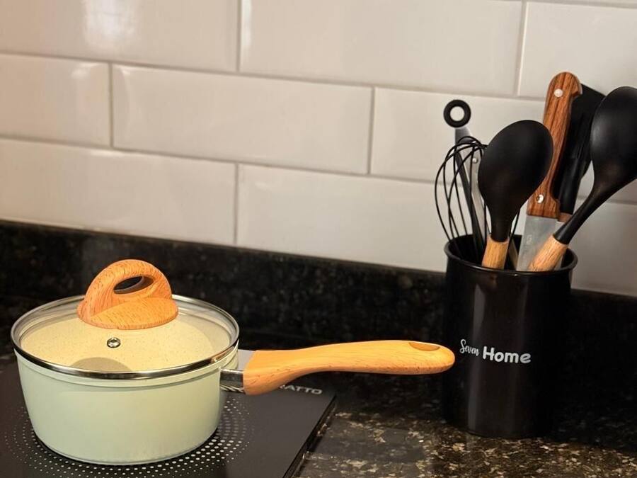 Una cocina con una olla de vidrio sobre una cocina de inducción, una cuchara de mango de madera y un portalapices con utensilios negros y de madera.