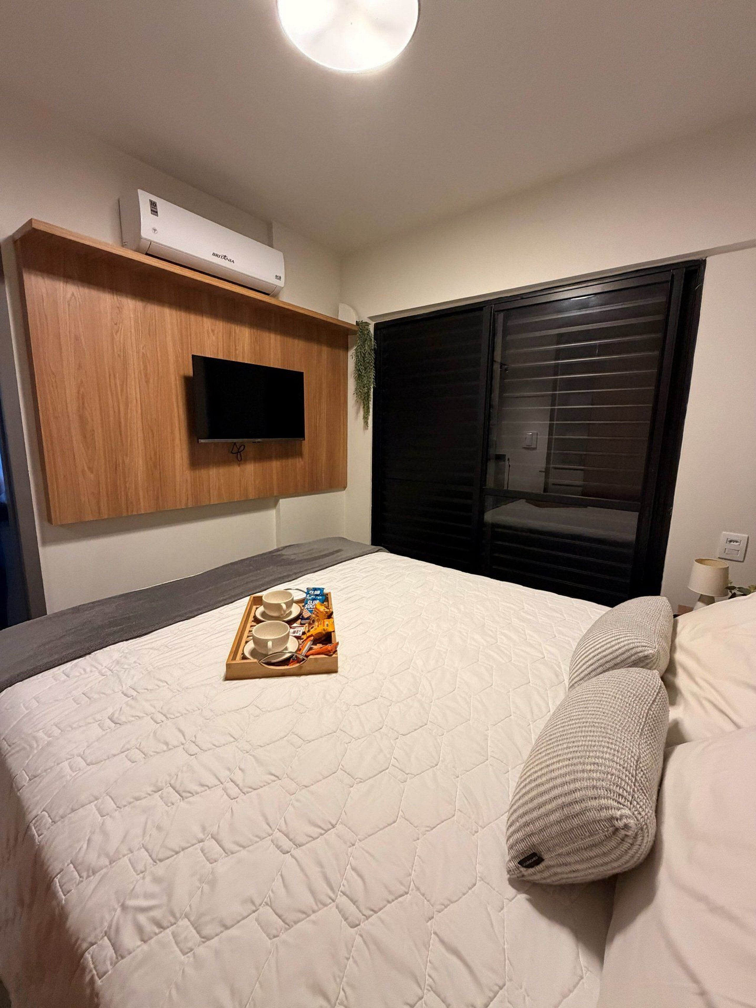 Quarto de hotel com uma cama, uma televisão de tela plana fixada em um painel de madeira, uma janela com persianas e uma bandeja com itens de café da manhã sobre a cama.