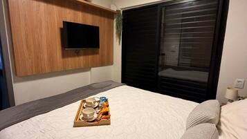 Quarto de hotel com uma cama, uma televisão de tela plana fixada em um painel de madeira, uma janela com persianas e uma bandeja com itens de café da manhã sobre a cama.