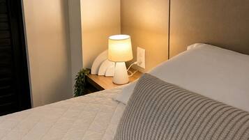 Uma cama bem arrumada com cabeceira acolchoada, luminária de mesa e uma pequena planta na mesinha de cabeceira.