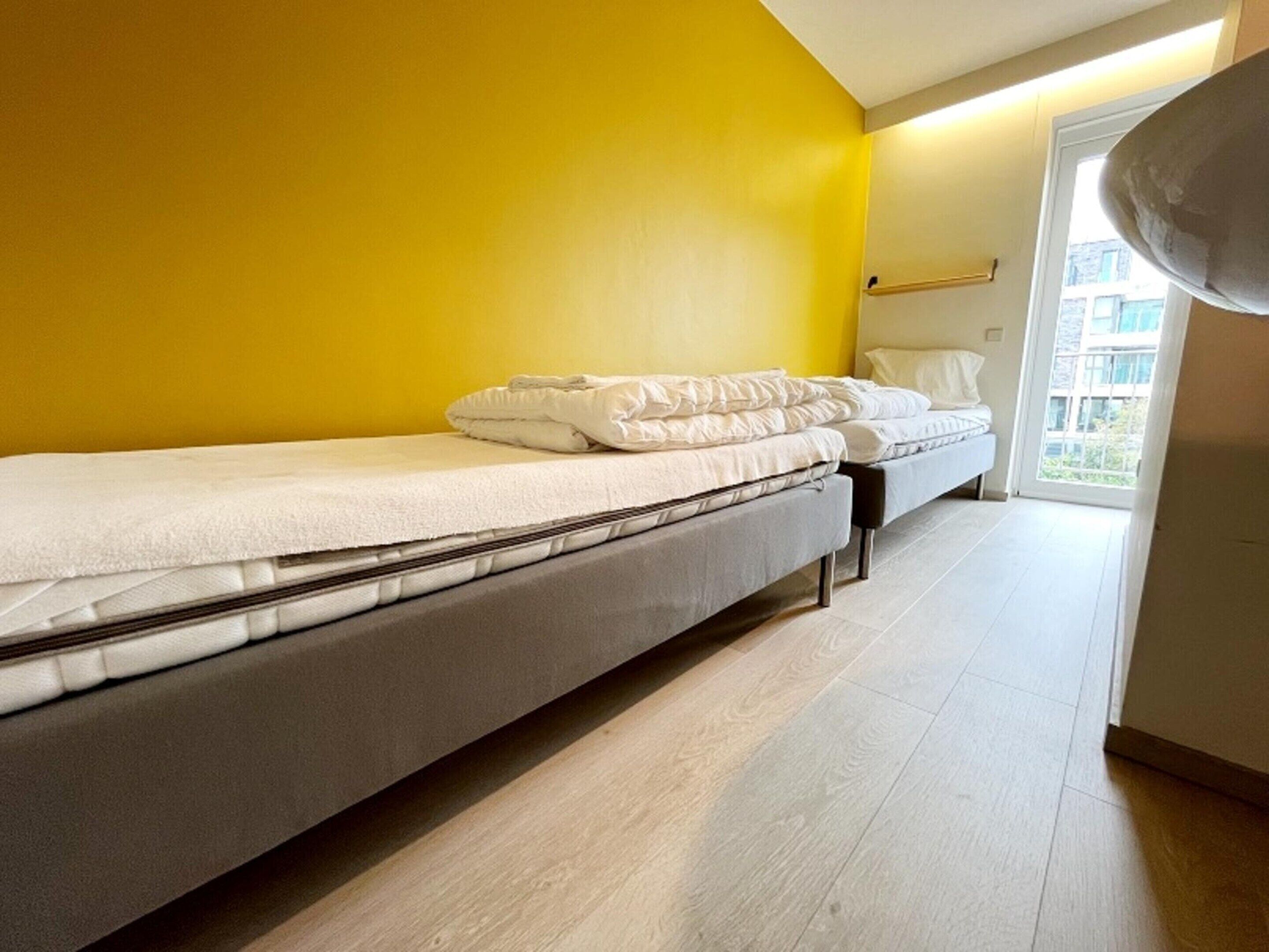 Une chambre avec deux lits, un mur jaune et une fenêtre donnant sur des immeubles.